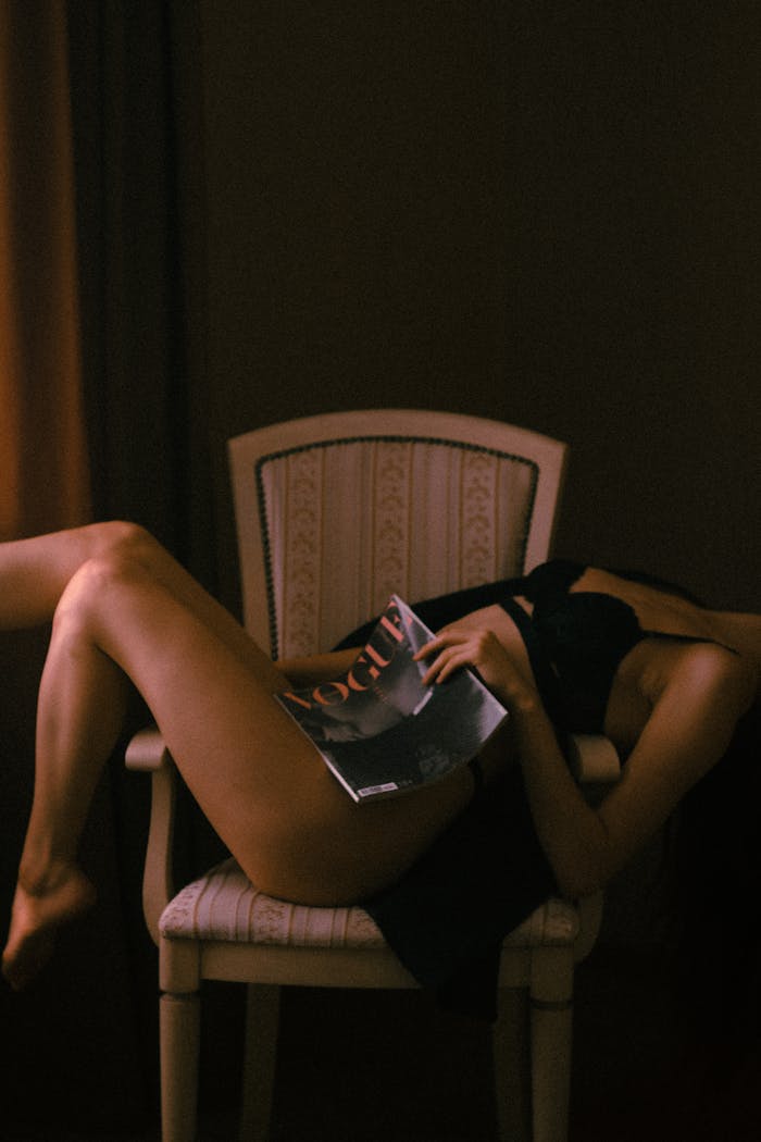 A woman in lingerie lounging on a chair, holding a fashion magazine, creating a sultry and alluring atmosphere.